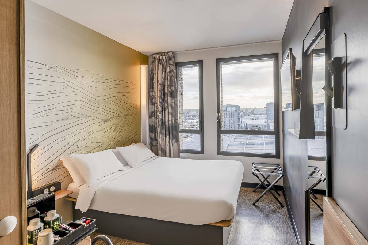 Ein Hotelzimmer mit Bett, einem kleinen Tisch mit Stuhl, einem Fenster mit Blick auf die Stadt und einem an der Wand befestigten Fernseher.