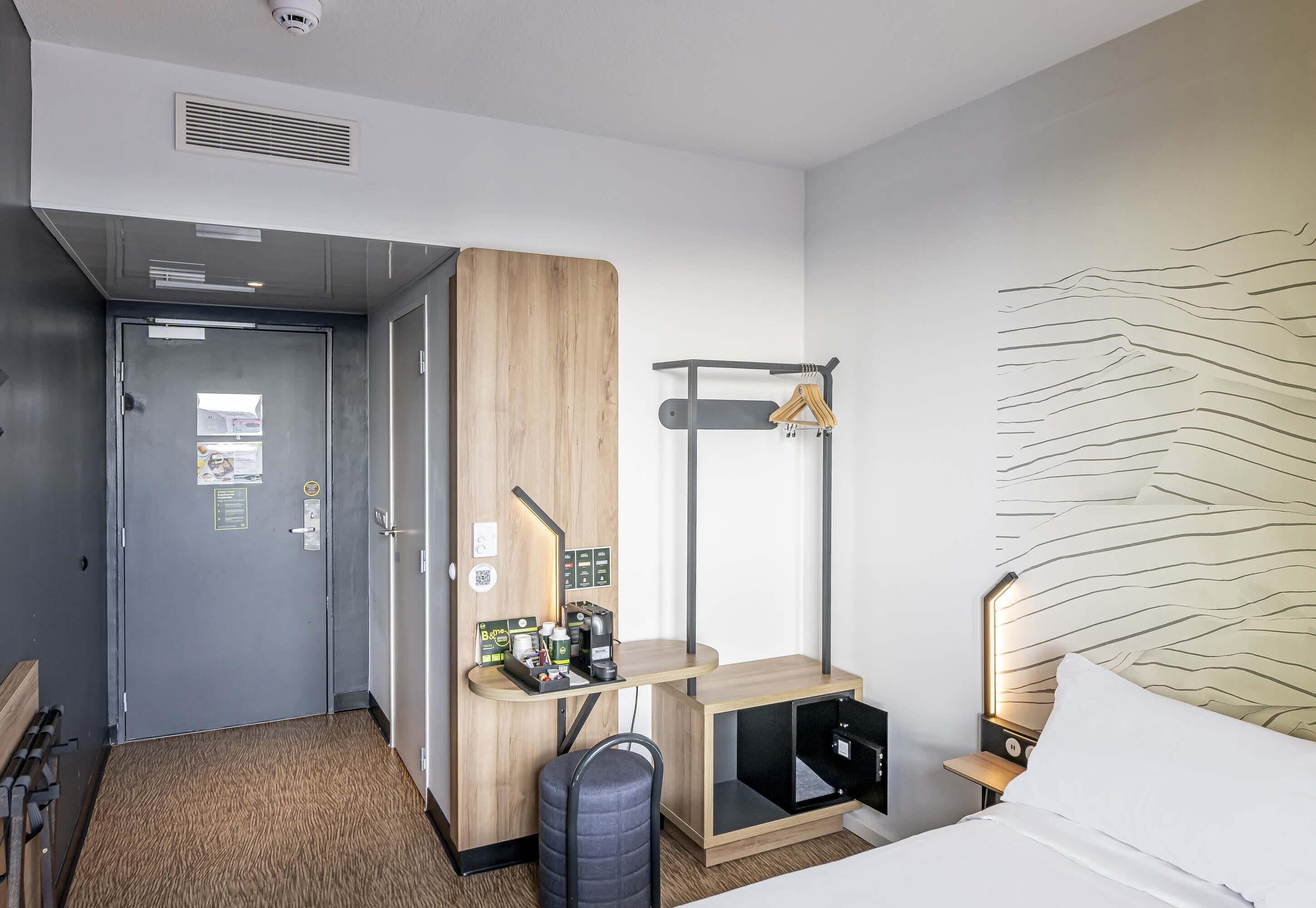 Une chambre d’hôtel comprenant un lit, un bureau avec une chaise, un porte-manteau, une lampe fixée au mur et une porte dotée d’une grille d’aération.