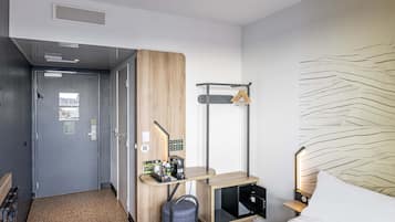 Une chambre d’hôtel comprenant un lit, un bureau avec une chaise, un porte-manteau, une lampe fixée au mur et une porte dotée d’une grille d’aération.