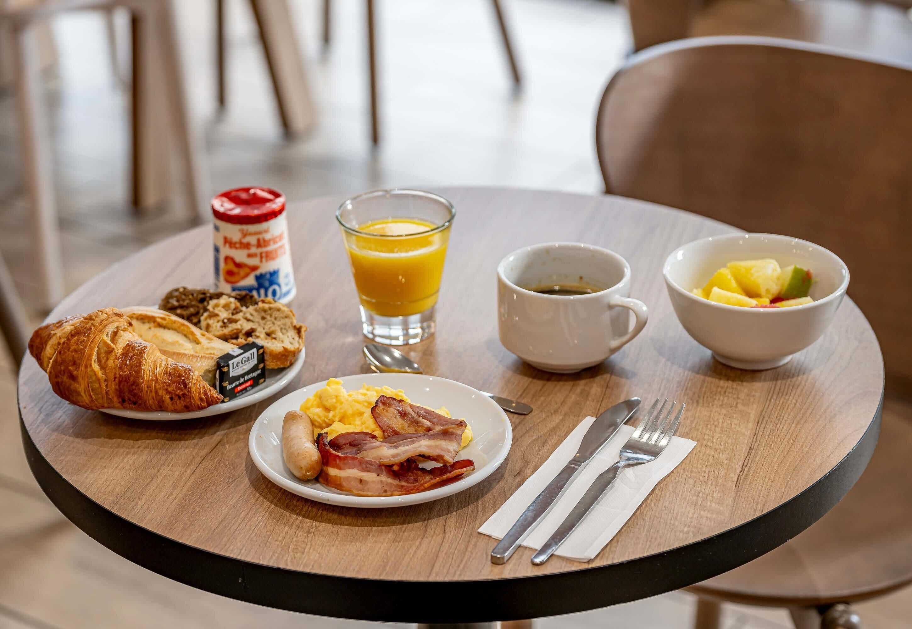 Una mesa de desayuno con croissants, huevos revueltos, tocino, zumo de naranja, café y un bol de fruta.