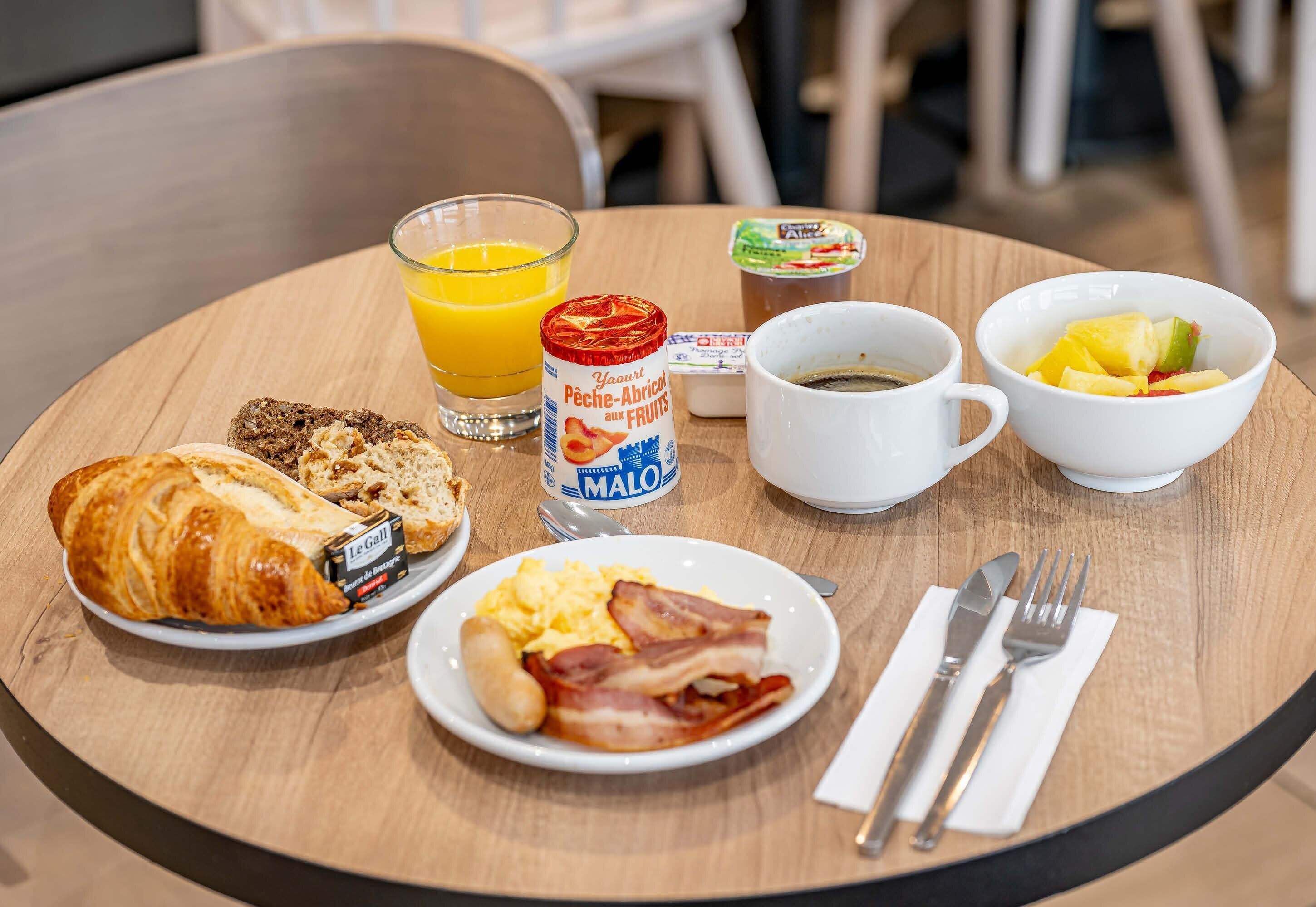 Desayuno con cruasanes, huevos revueltos, tocino, un vaso de jugo de naranja, yogur y un tazón de fruta sobre una mesa de madera.