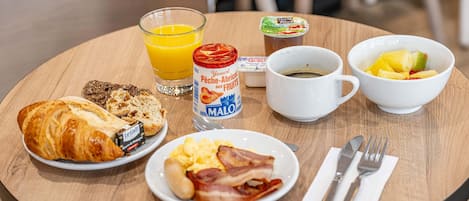 A breakfast setting with croissants, scrambled eggs, bacon, a glass of orange juice, yogurt, and a bowl of fruit on a wooden table.