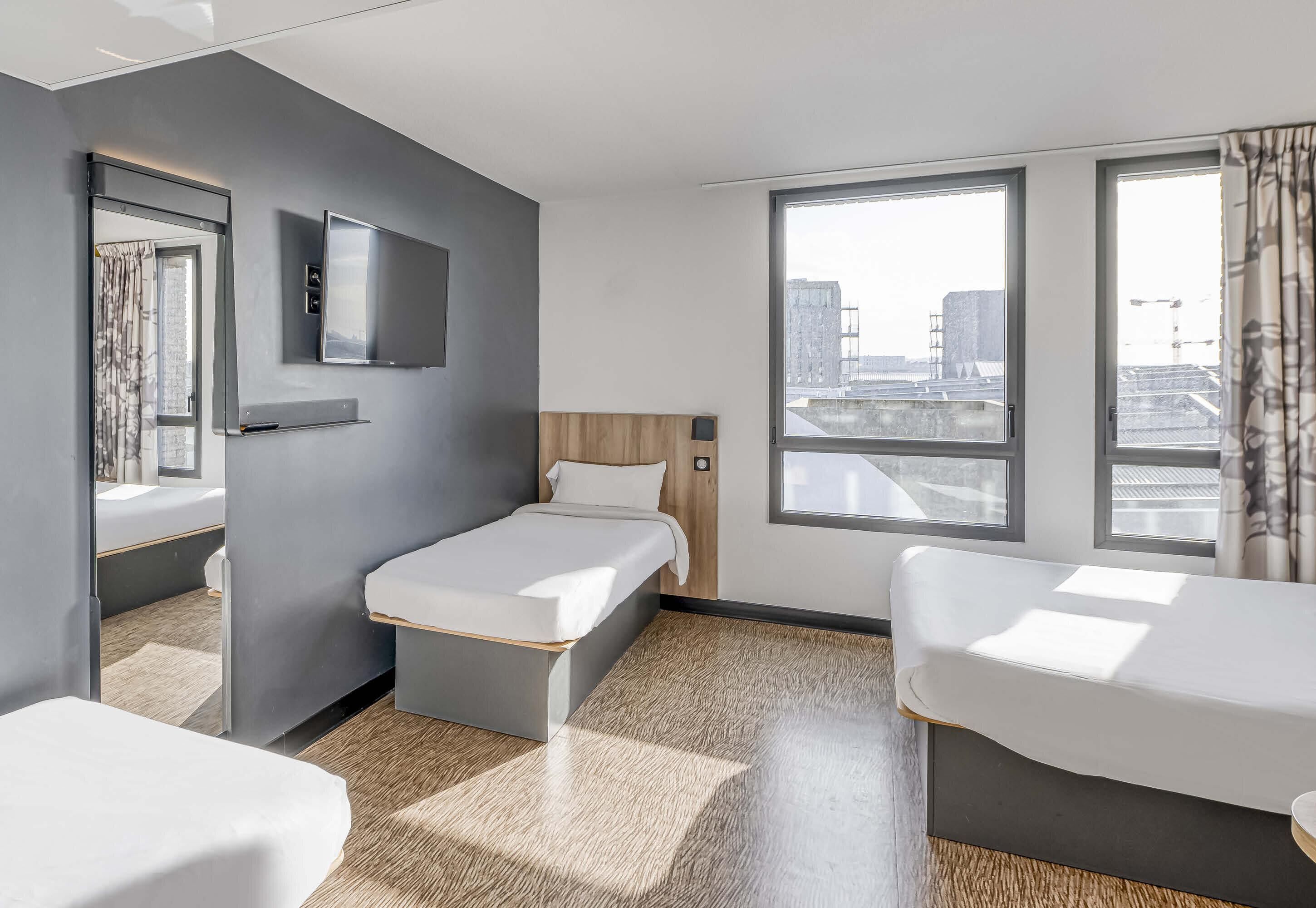 Quarto de hotel com cama, escrivaninha, televisão e amplas janelas com vista para a cidade.