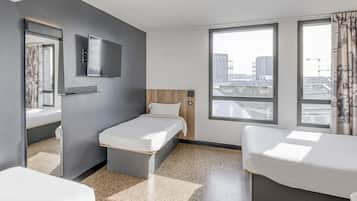 Quarto de hotel com cama, escrivaninha, televisão e amplas janelas com vista para a cidade.