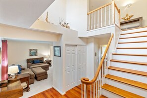 Un vaste espace de vie avec un escalier menant à l’étage supérieur, un coin salon confortable avec un canapé et un pouf, et une porte agrémentée d’un miroir et de cadres de photos.