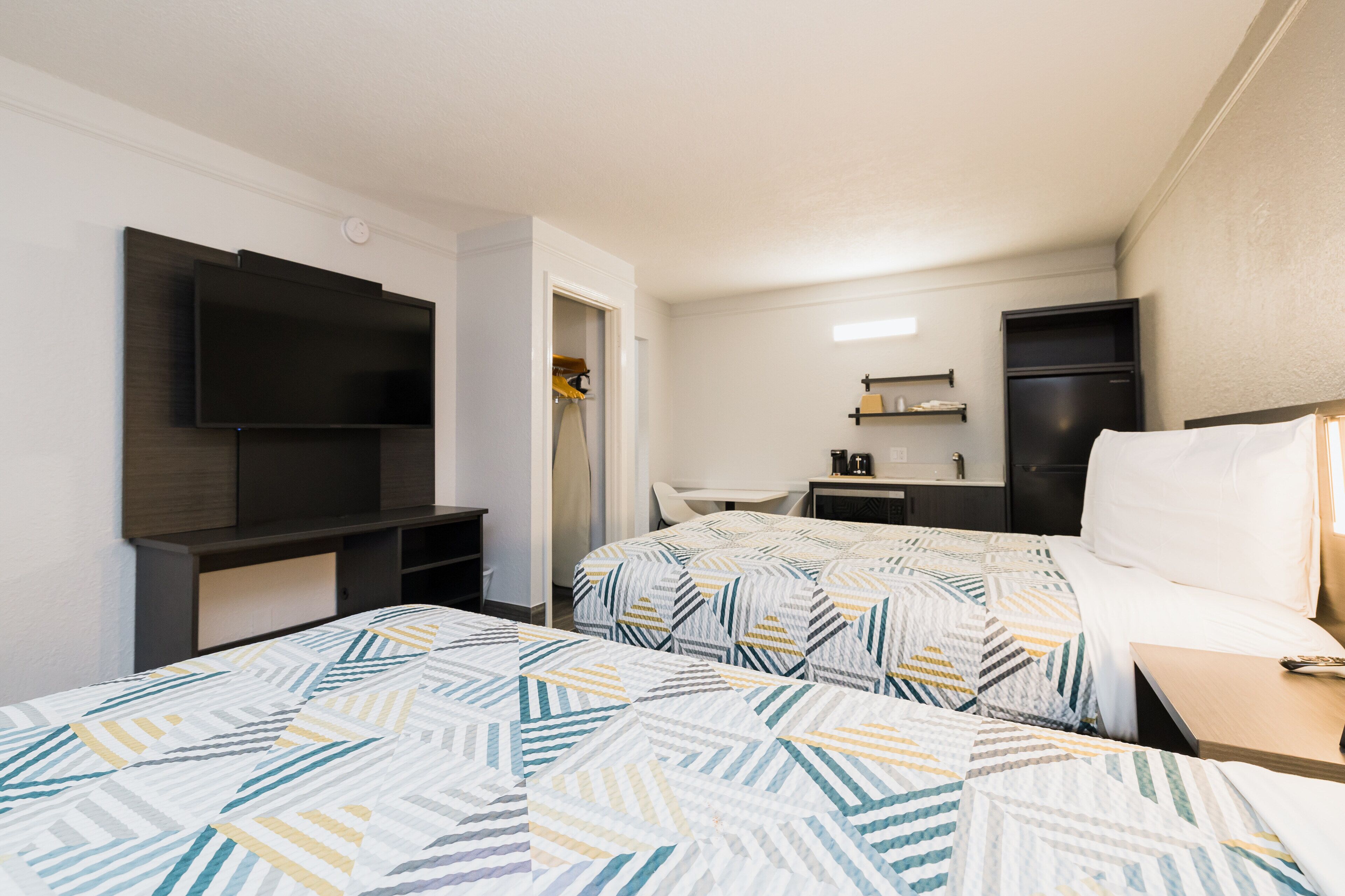 A hotel room with two beds, a TV, a desk, a refrigerator, and a small kitchenette.