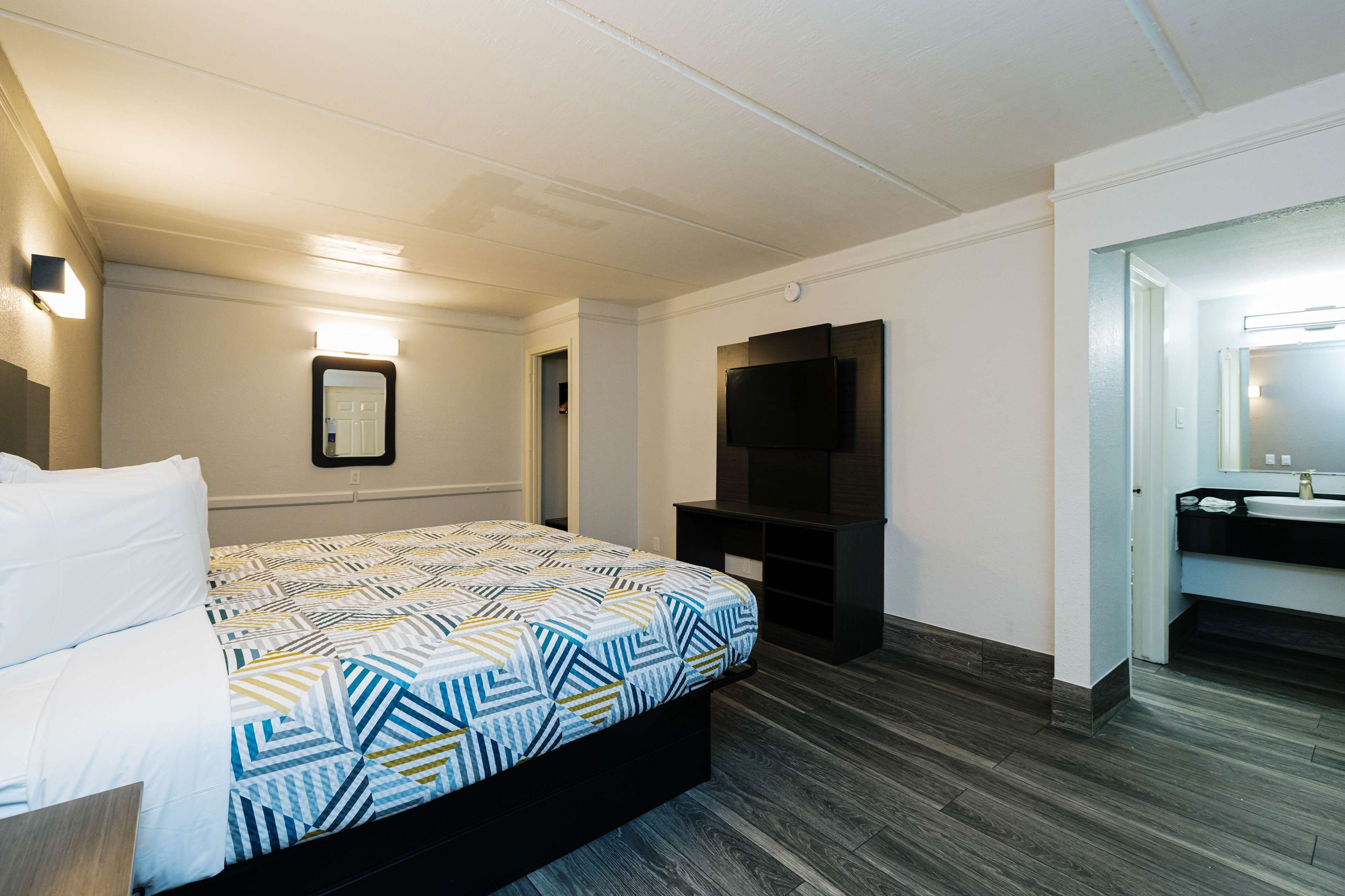 A hotel room with a bed, a TV, a mirror, and a bathroom with a sink.