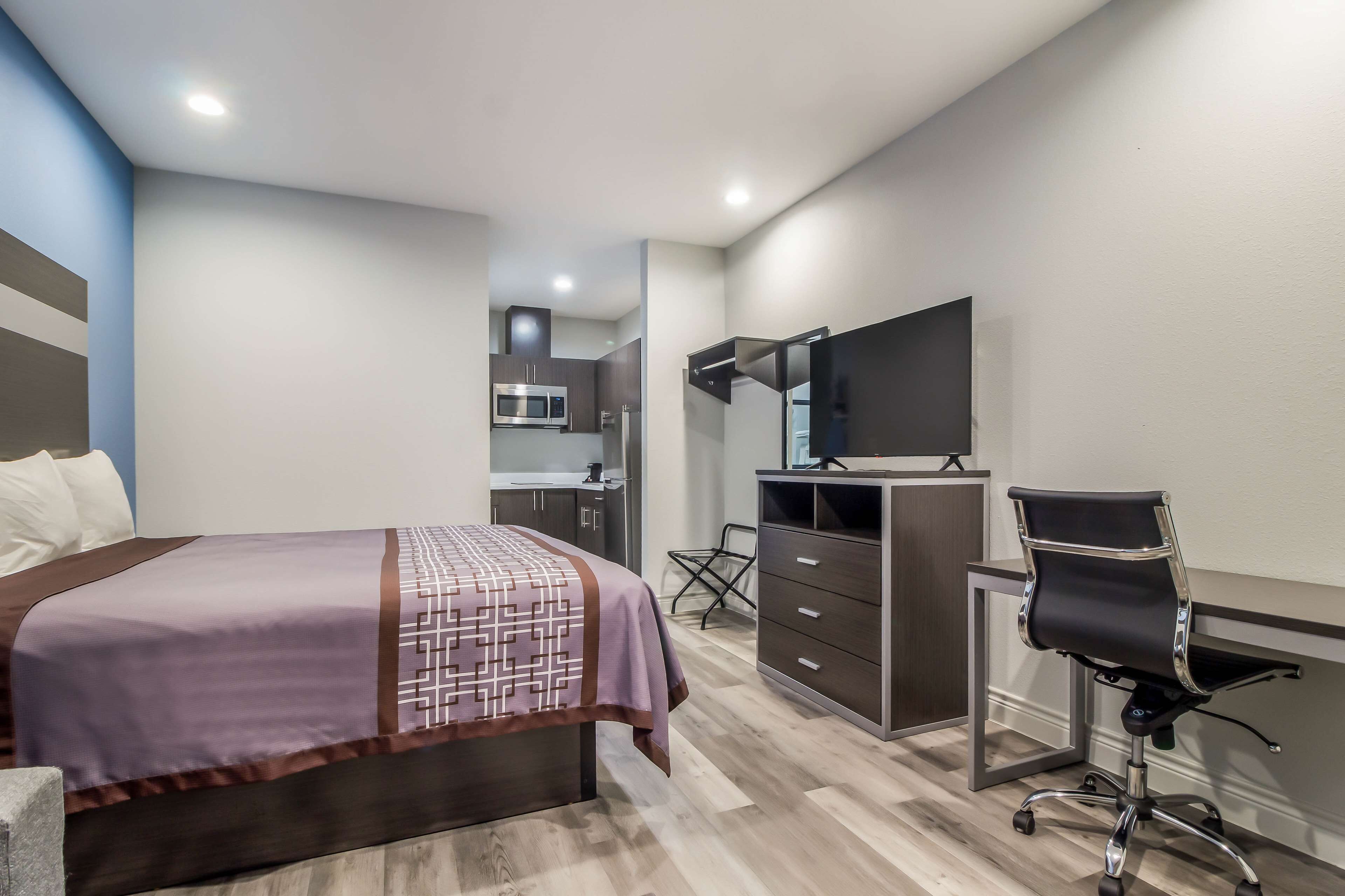 A modern hotel room with a bed, a desk, a chair, a flat-screen TV, and a kitchenette in the background.