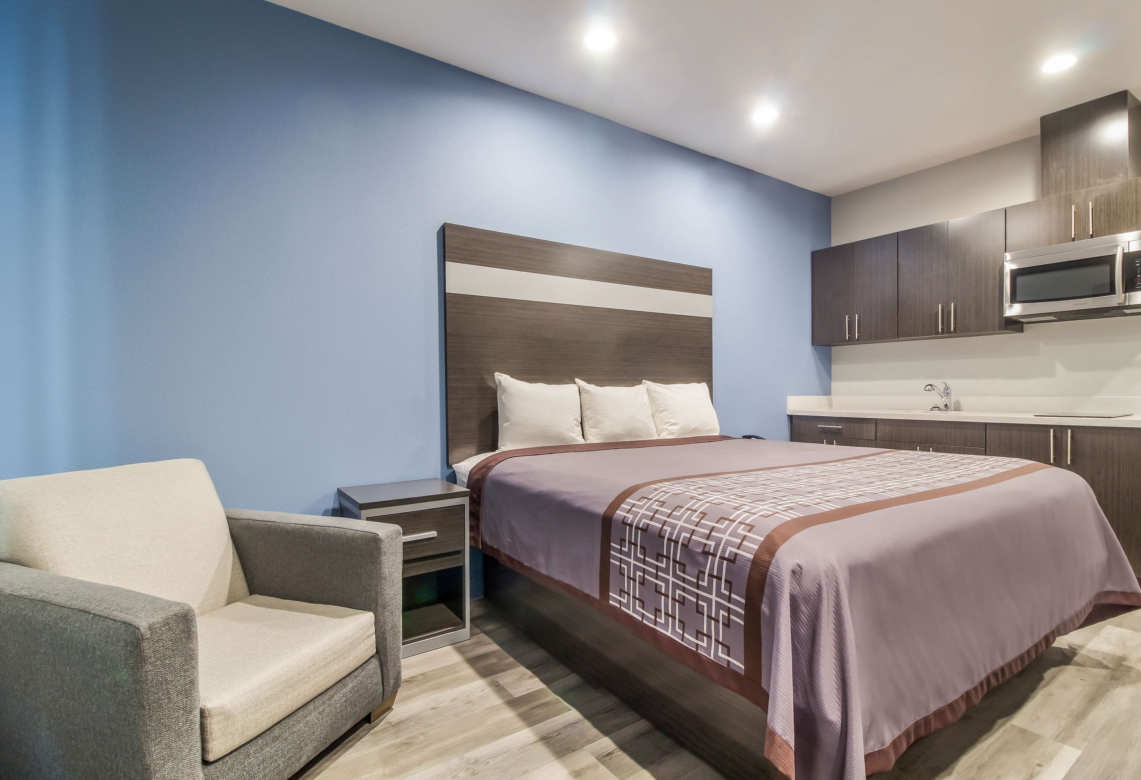Una habitación de hotel moderna con una cama, una silla, una mesita de noche, un microondas y un lavamanos.