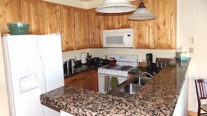 Eine Küche mit Holzausstattung, einem weißen Kühlschrank, einer Mikrowelle, einem Herd, Granit-Arbeitsplatten und hängenden Pendelleuchten.