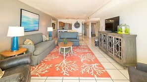 Uma sala de estar com sofá, mesa de centro e um tapete vermelho estampado. Há uma televisão e um armário com um objeto decorativo em cima.