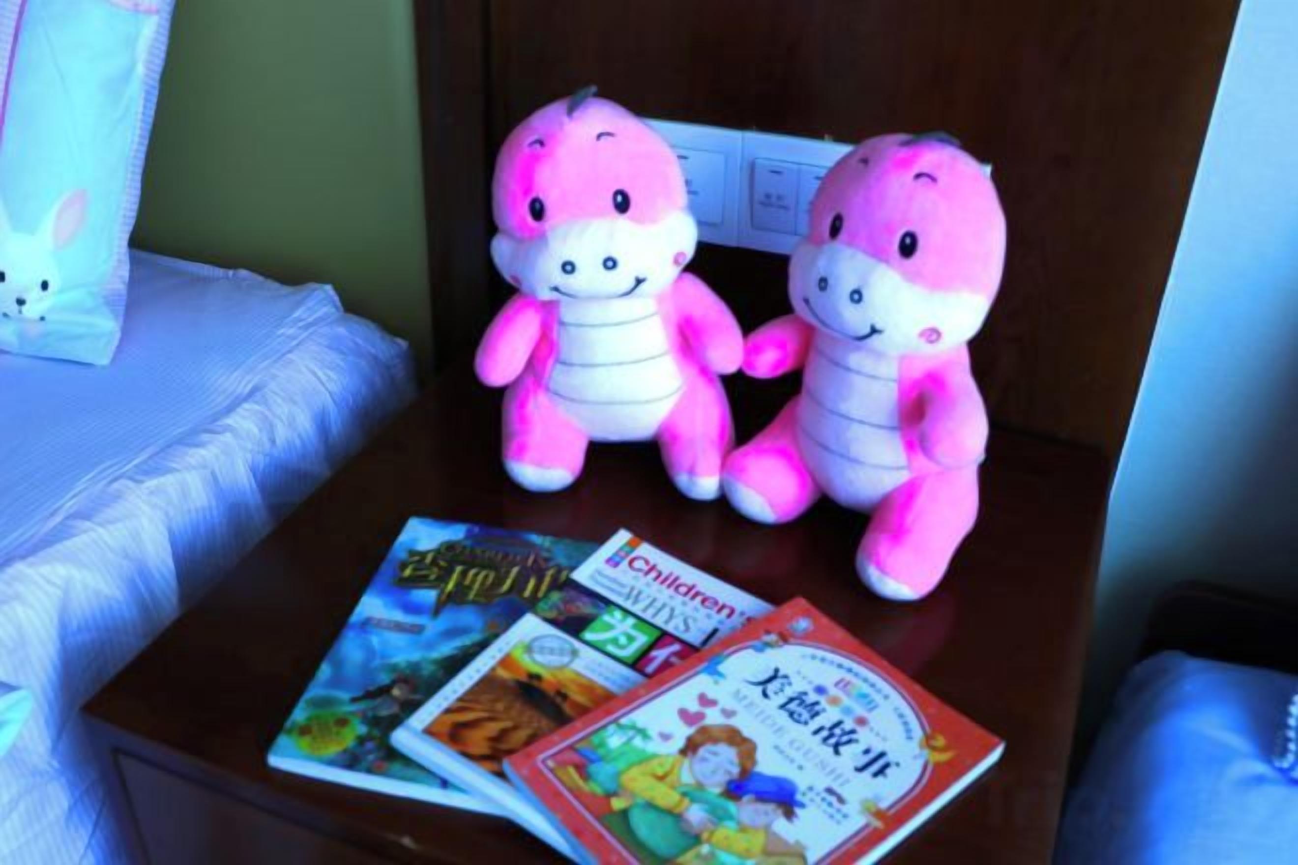 A bed with a white comforter and pillows, a wooden nightstand with two plush toys and children's books.