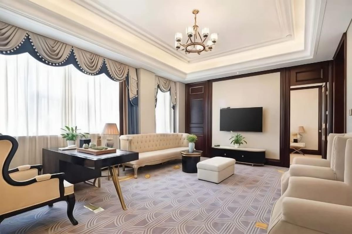 Quarto de hotel com sofá, escrivaninha, televisão e lustre.