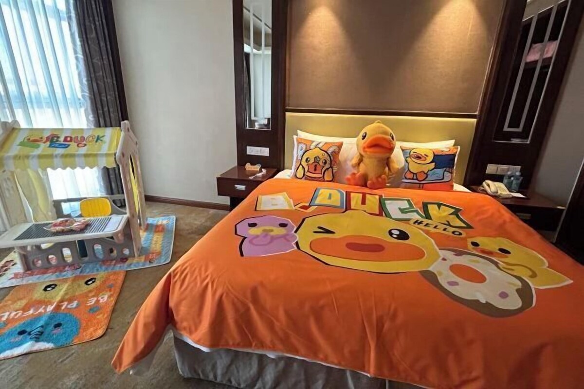 A hotel room with a bed featuring a colorful bedspread with cartoon duck characters. There is a small table with a toy duck and a chair.