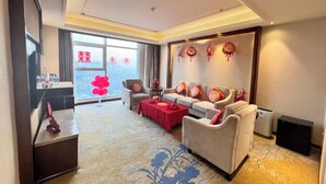 A living room with a sofa, a coffee table, and a TV. There is a window with curtains and a view of the city. - Blue Horizon Jun Hua Hotel (Fei County)