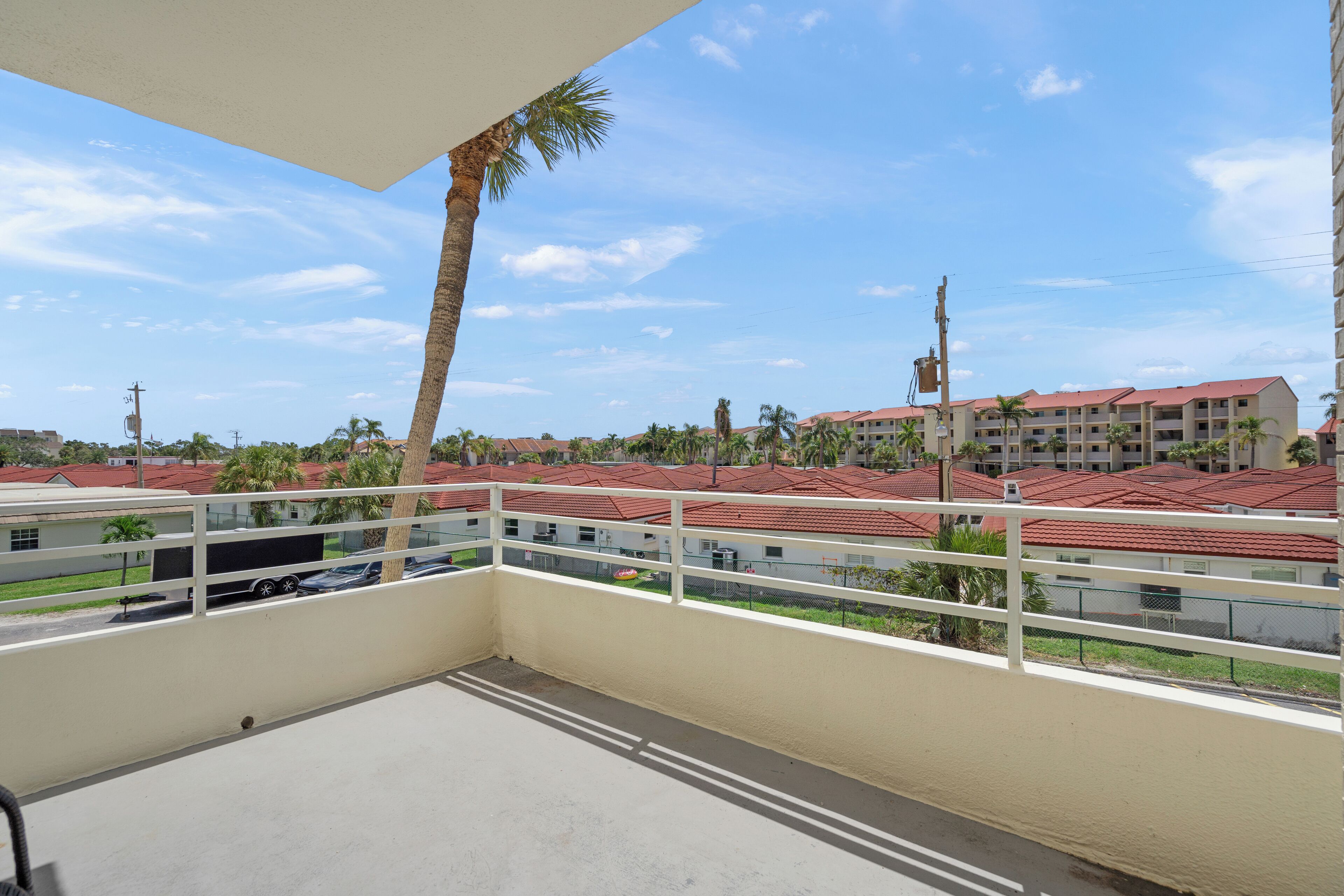 A balcony with a view of a residential area, including buildings with red roofs and palm trees.