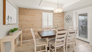 Un coin repas avec une table et des chaises en bois, un luminaire suspendu, une horloge au mur et une fenêtre avec des volets.
