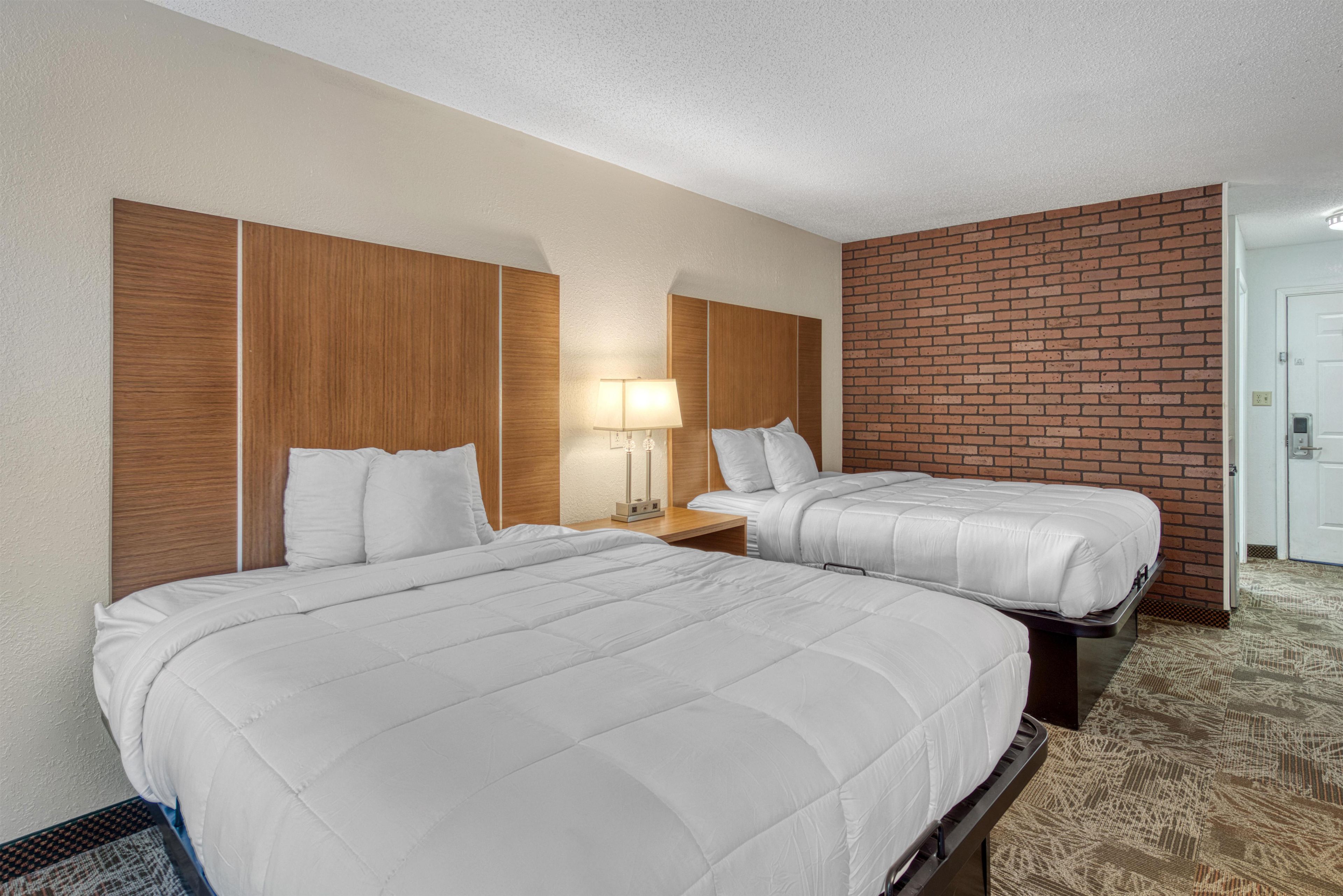 A hotel room with two beds, a wooden headboard, a brick accent wall, and a carpeted floor.