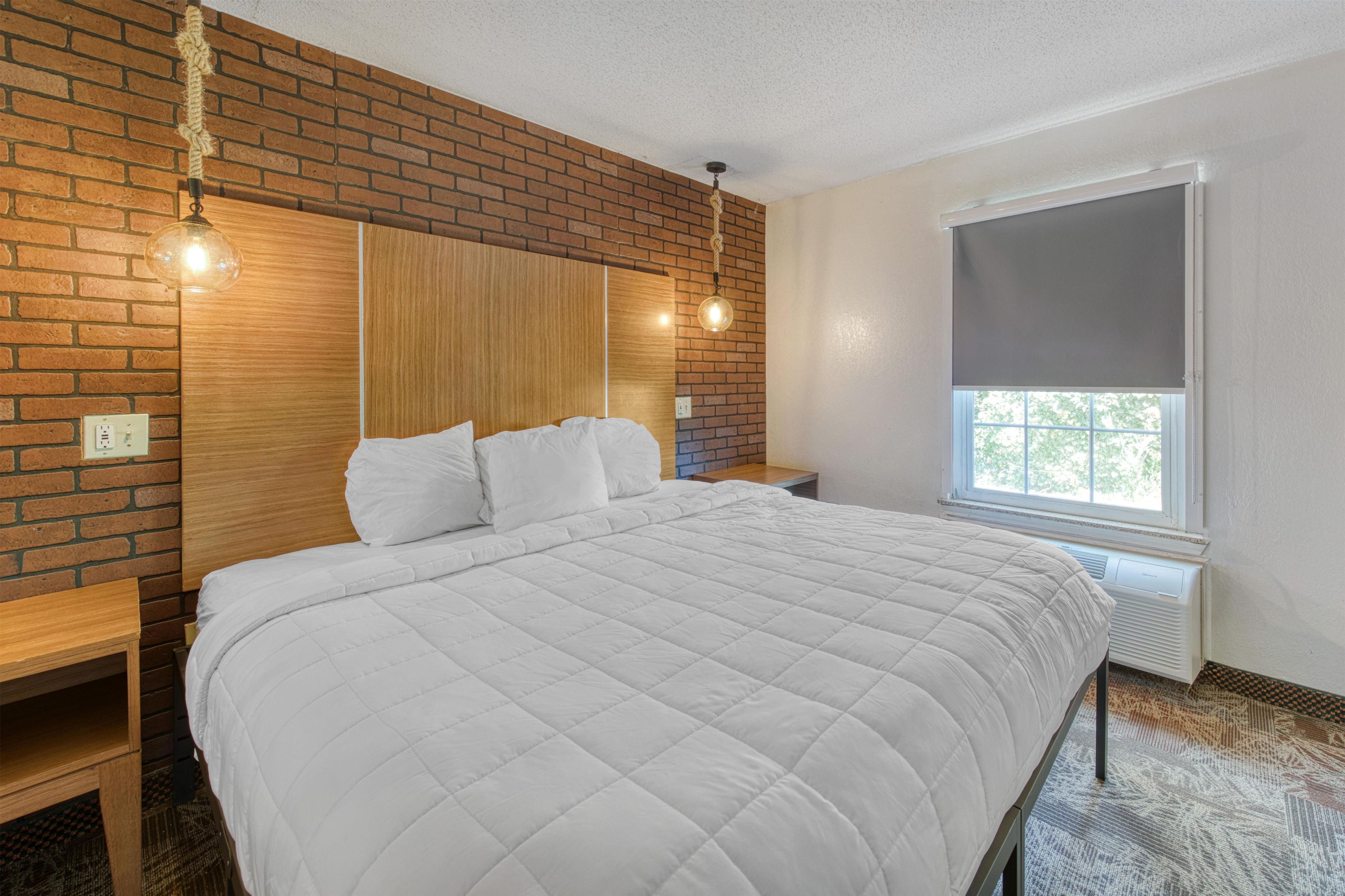 A bedroom with a brick wall, a wooden headboard, a large bed with white bedding, a bedside table, a hanging light fixture, and a window with a blind.