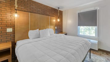 A bedroom with a brick wall, a wooden headboard, a large bed with white bedding, a bedside table, a hanging light fixture, and a window with a blind.