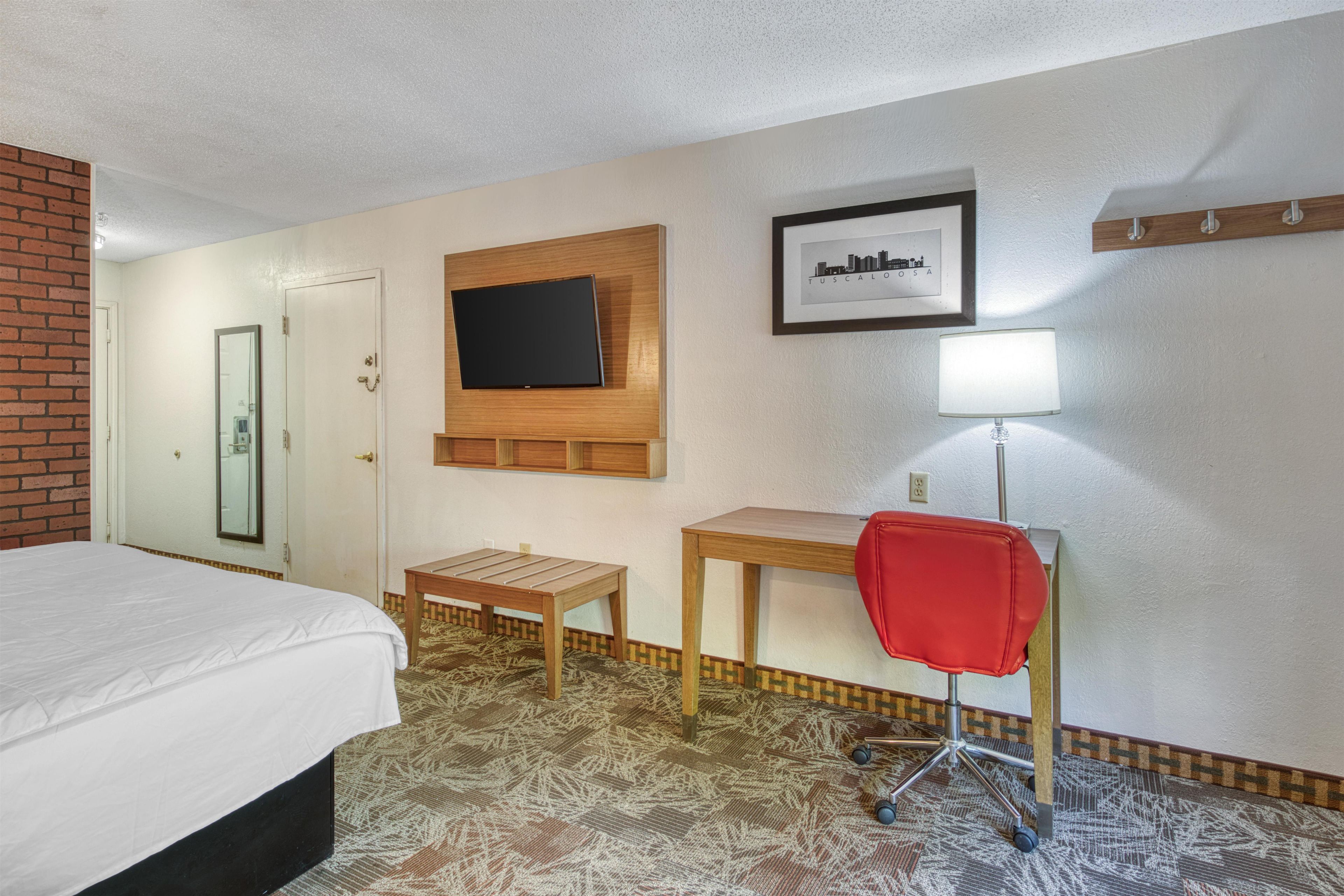 A hotel room with a bed, a television mounted on the wall, a small wooden table, a red office chair, and a framed picture on the wall.