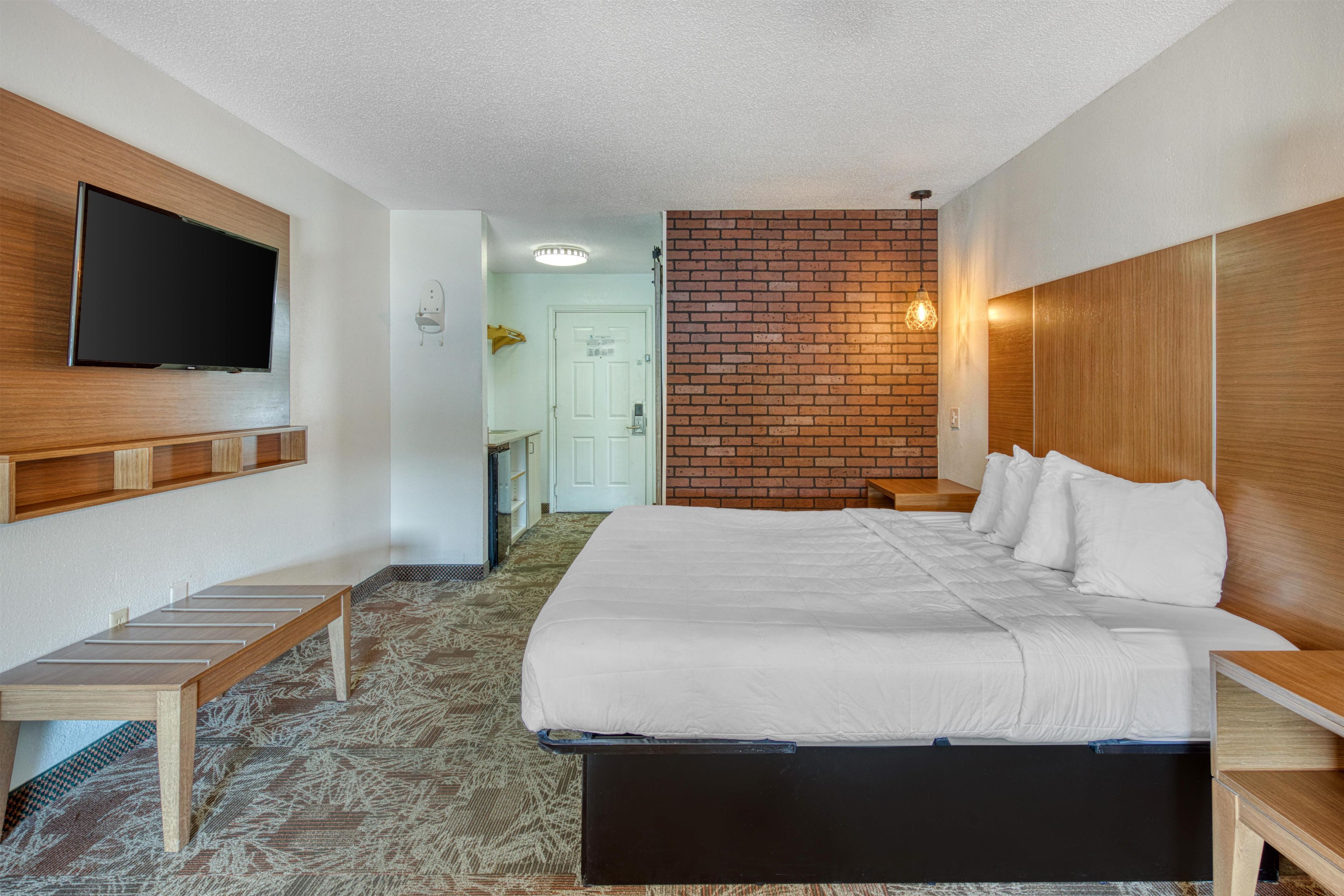 A hotel room with a bed, a TV mounted on the wall, a wooden bench, and a brick accent wall.