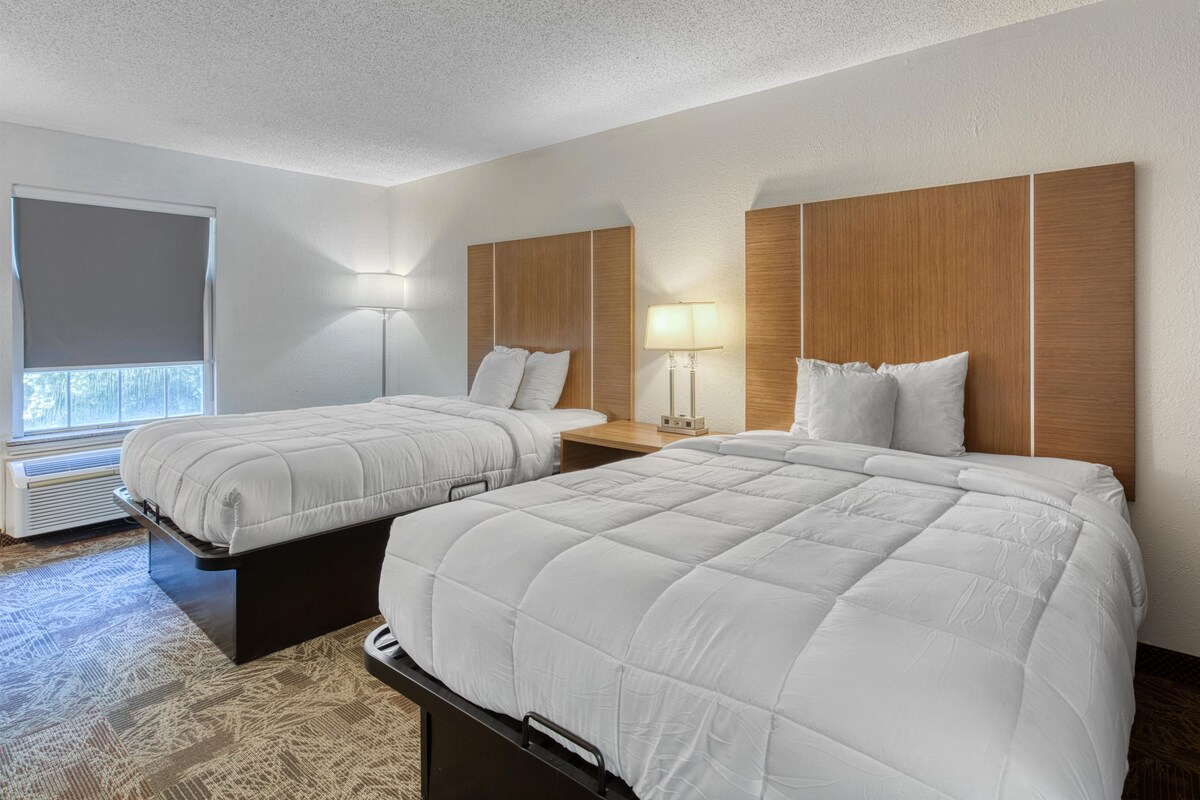 A hotel room with two beds, a window with a view, a lamp, and a heater.