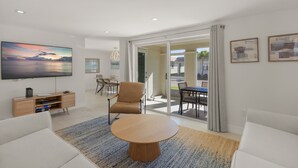 Un salon moderne avec un téléviseur à écran plat, une table basse ronde en bois, un fauteuil beige et un canapé. On aperçoit un coin repas avec une table et des chaises à travers une porte vitrée.