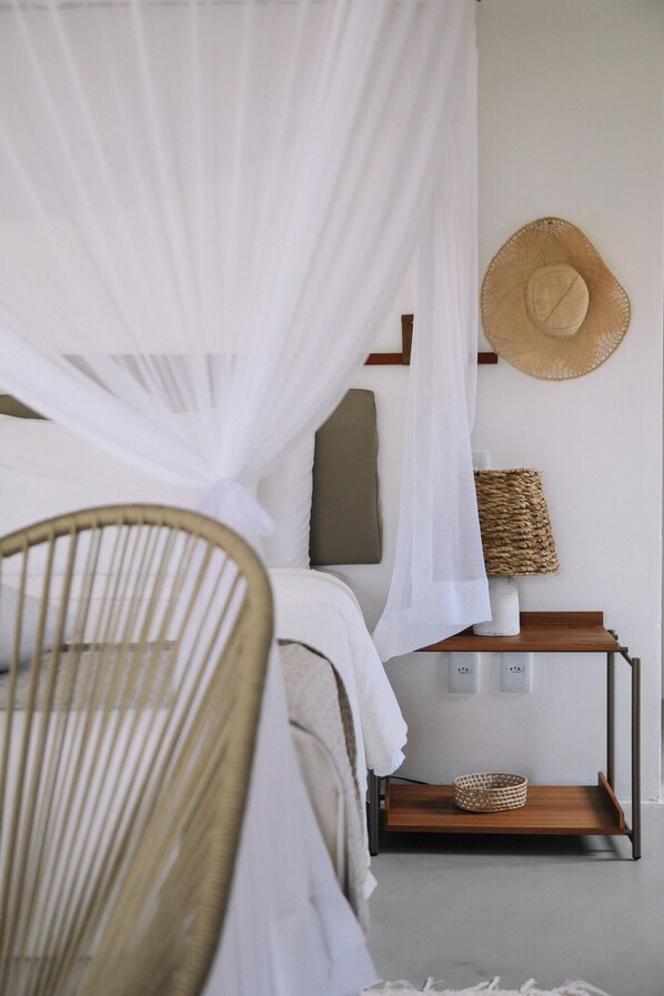 A bedroom with a bed under a white canopy, a wicker chair, a woven basket, and a straw hat on the wall.