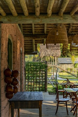 An outdoor dining area with wooden tables and chairs, a thatched roof, and a view of a garden and building in the background. - Villa dos Nativos Boutique Hotel (Porto Seguro)