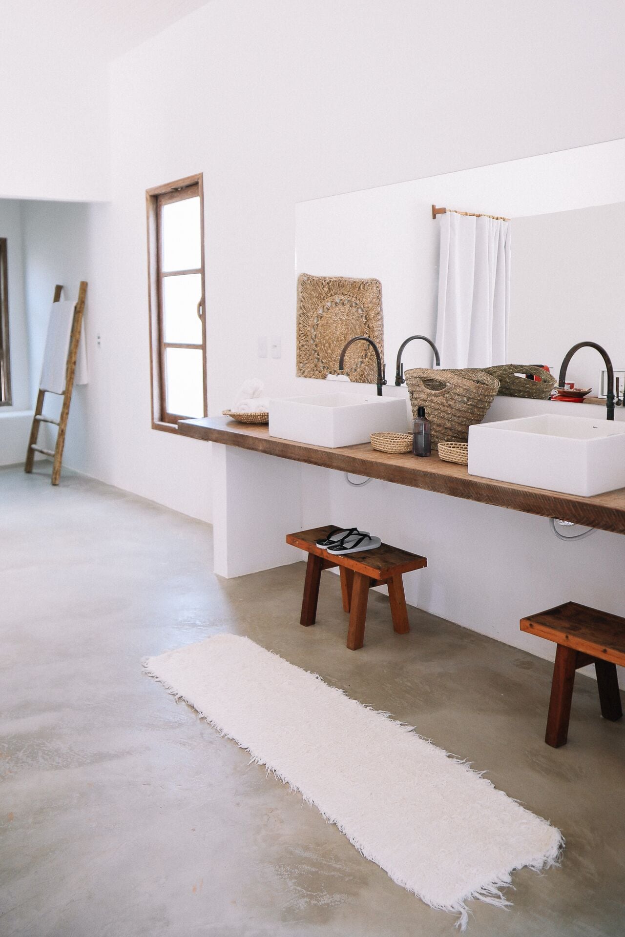 Un baño minimalista con un espejo grande, dos lavamanos, un taburete de madera y una alfombra blanca.