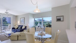 A spacious living area with a glass dining table, comfortable seating, a ceiling fan, and a view of the city through large windows.