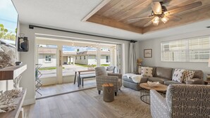 Ein geräumiges Wohnzimmer mit Holzbalkendecke, einem großen Fenster mit Jalousien, einem grauen sofa mit Muster und einem Essbereich mit Tisch und Stühlen.