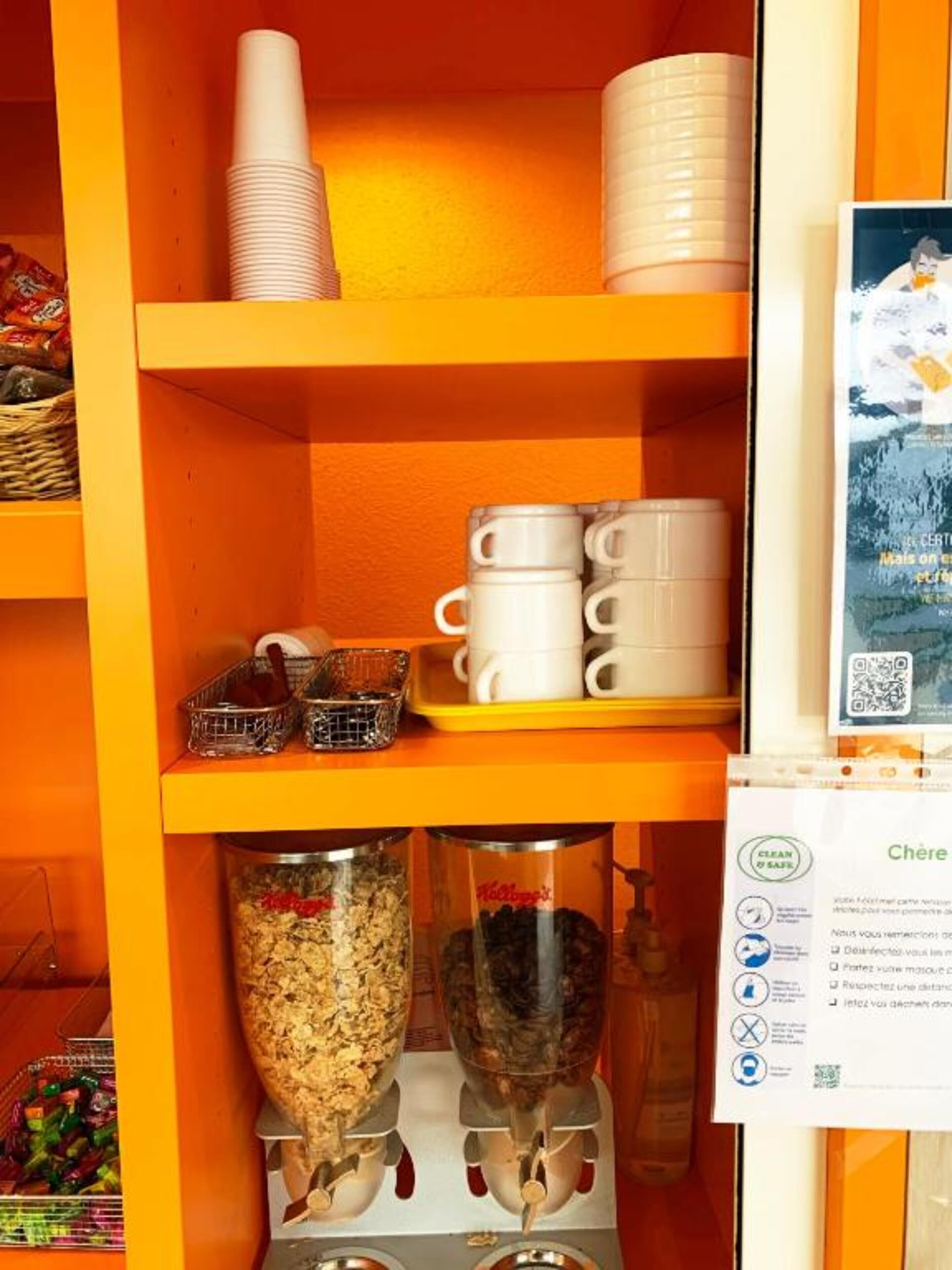 Uma prateleira com copos brancos empilhados, uma bandeja com canecas brancas e um dispensador de alimentos com um recipiente de cereal e um pote de nozes.