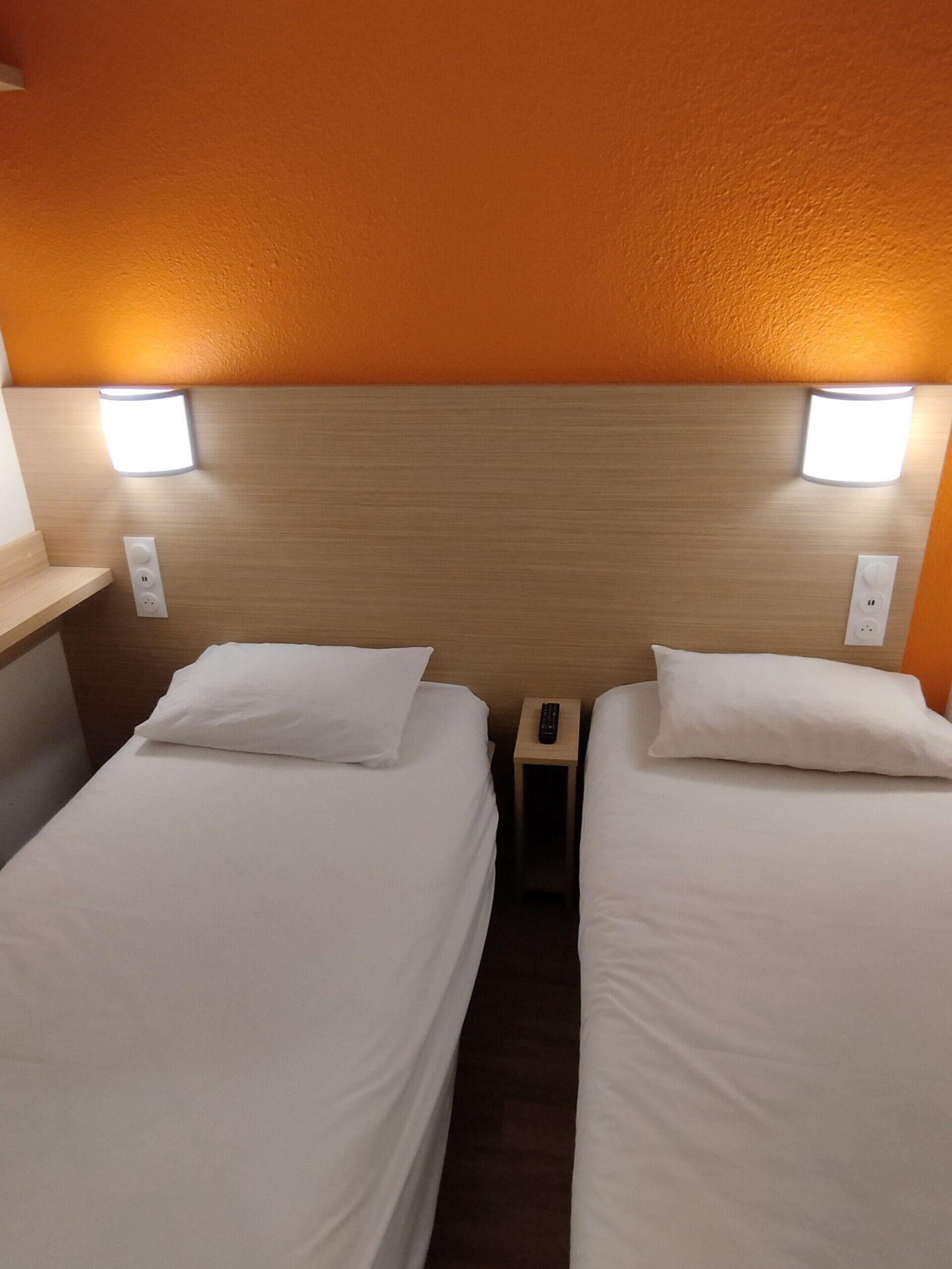 Habitación de hotel con dos camas individuales, cada una con una almohada blanca y una mesita de noche con un control remoto.