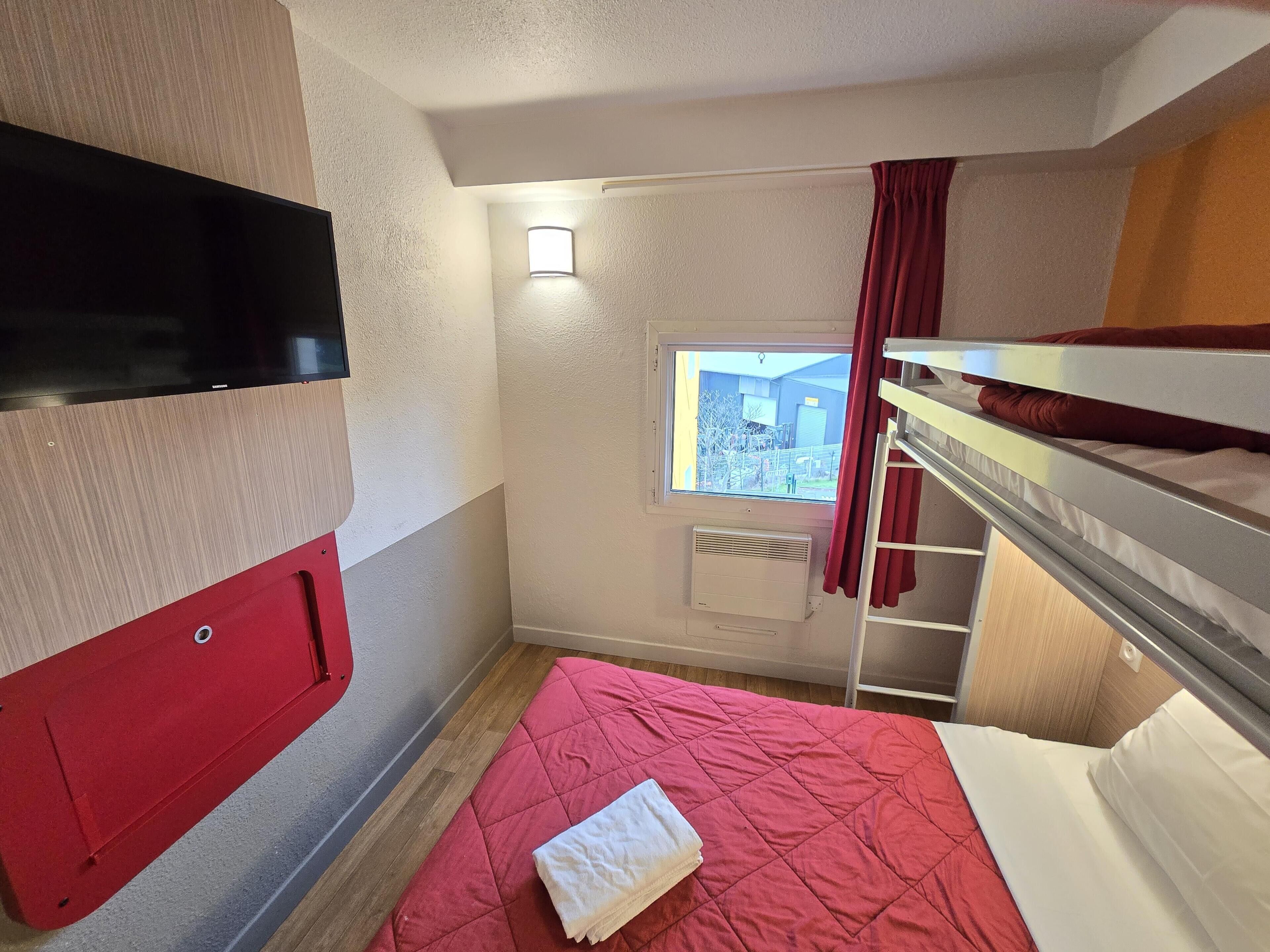 Een compacte hotelkamer met een stapelbed, een klein raam met gordijnen, een flatscreen televisie aan de muur en een rode sprei.
