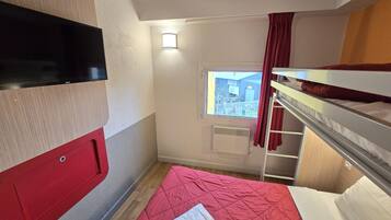 Een compacte hotelkamer met een stapelbed, een klein raam met gordijnen, een flatscreen televisie aan de muur en een rode sprei.