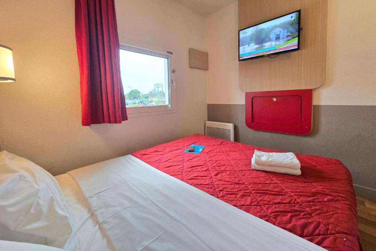 A hotel room with a bed, a TV mounted on the wall, a window with curtains, and a view of a building outside.
