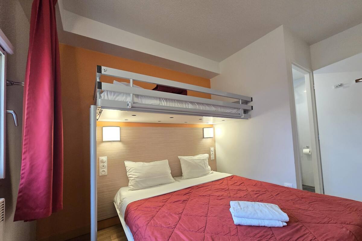 A single bed with a red quilt, a bunk bed above, and a red curtain on the left.