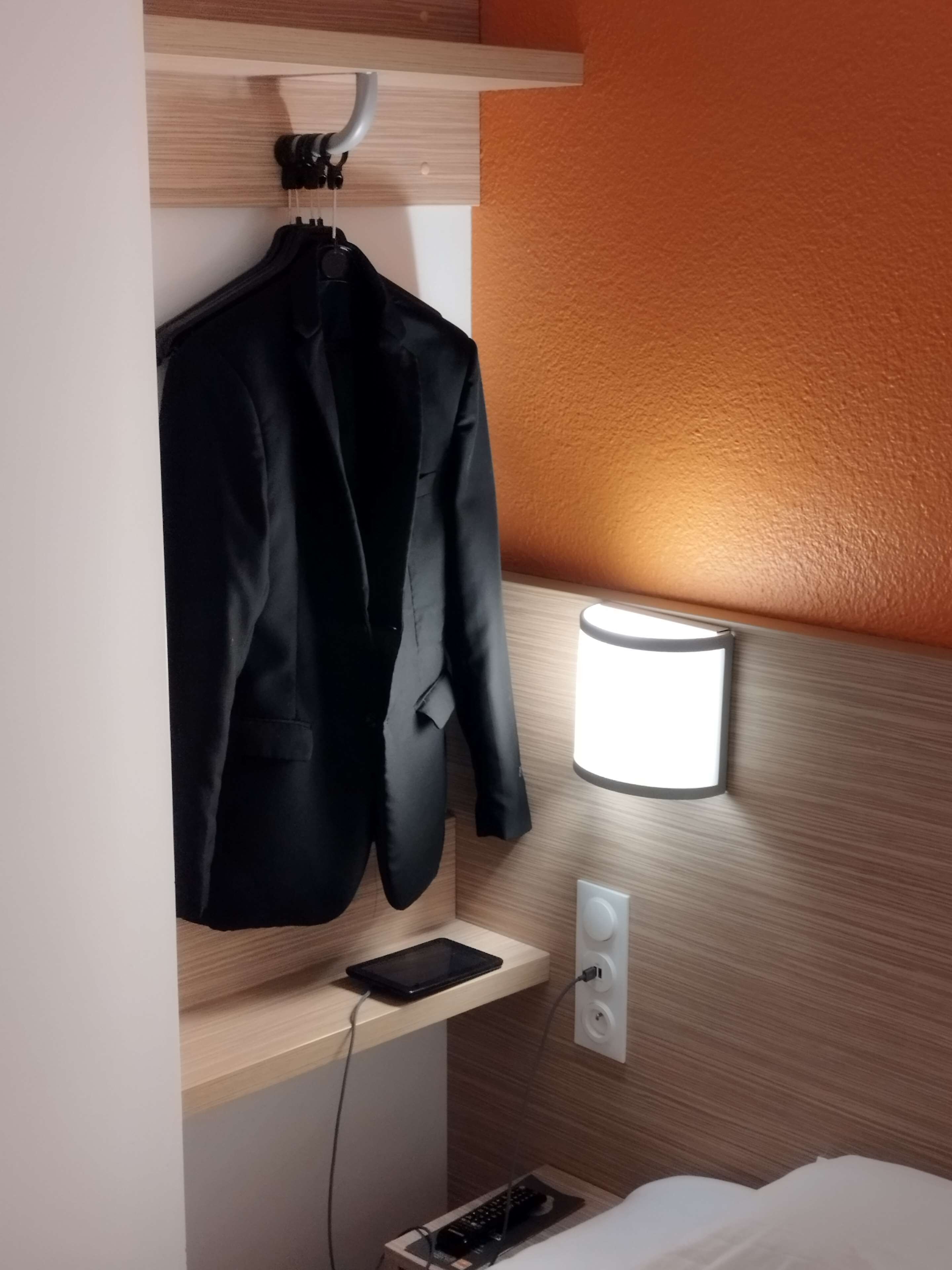 A hotel room with a wooden headboard, a bedside table with a lamp, a black jacket hanging on a hook, and a tablet on the table.