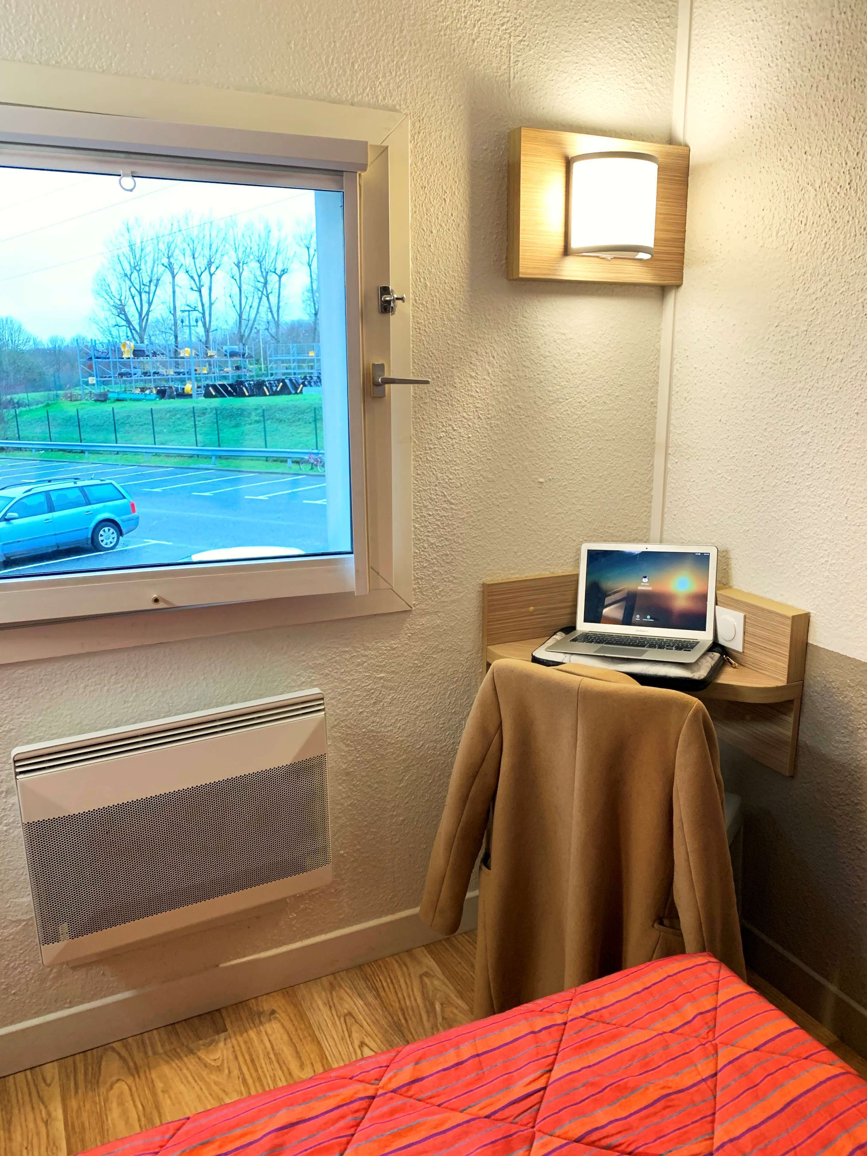 A hotel room with a window, a laptop on a small table, a radiator, and a bed with a red-striped blanket.