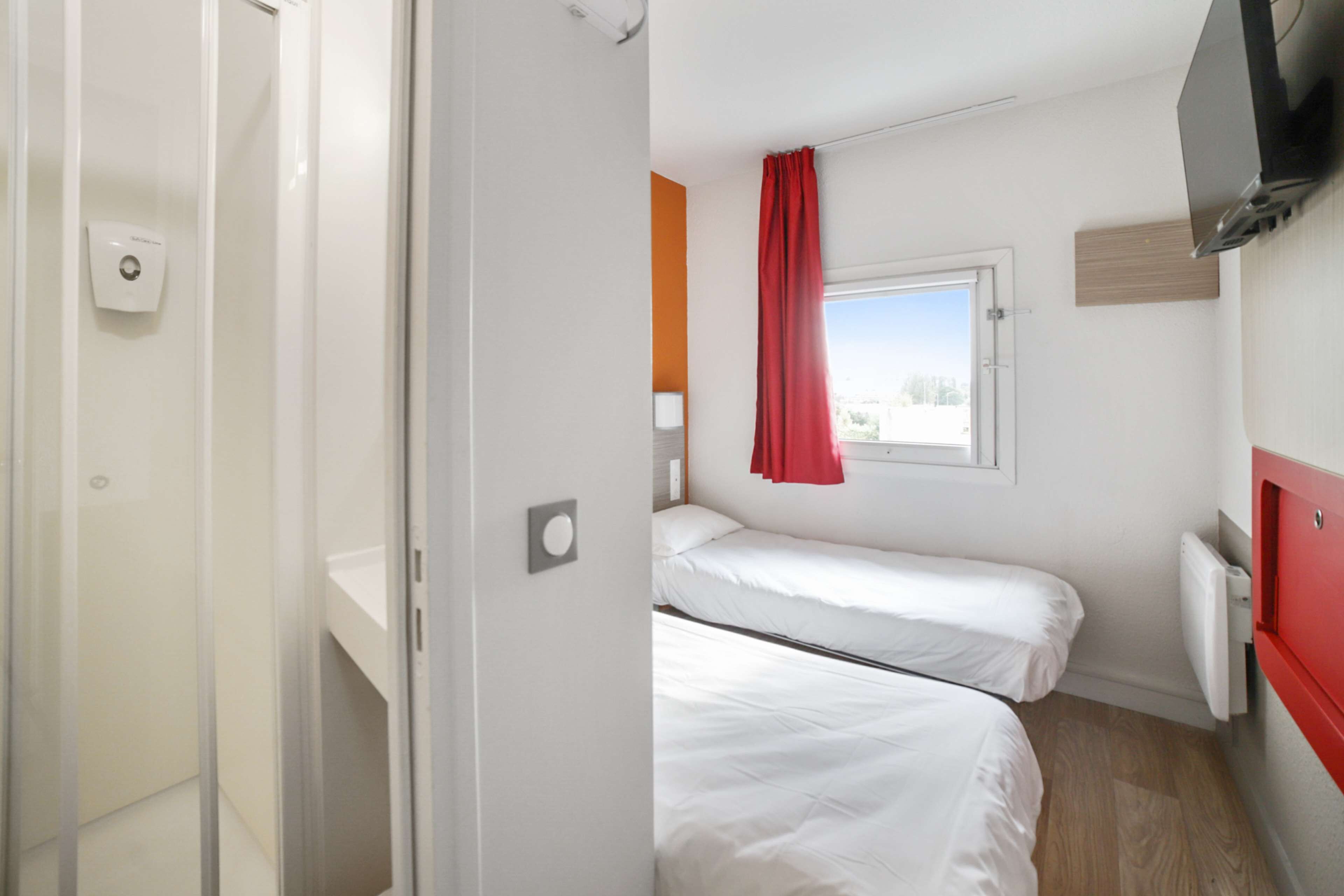 Une chambre avec deux lits simples, une fenêtre avec un rideau rouge, un téléviseur à écran plat fixé au mur et une porte blanche.