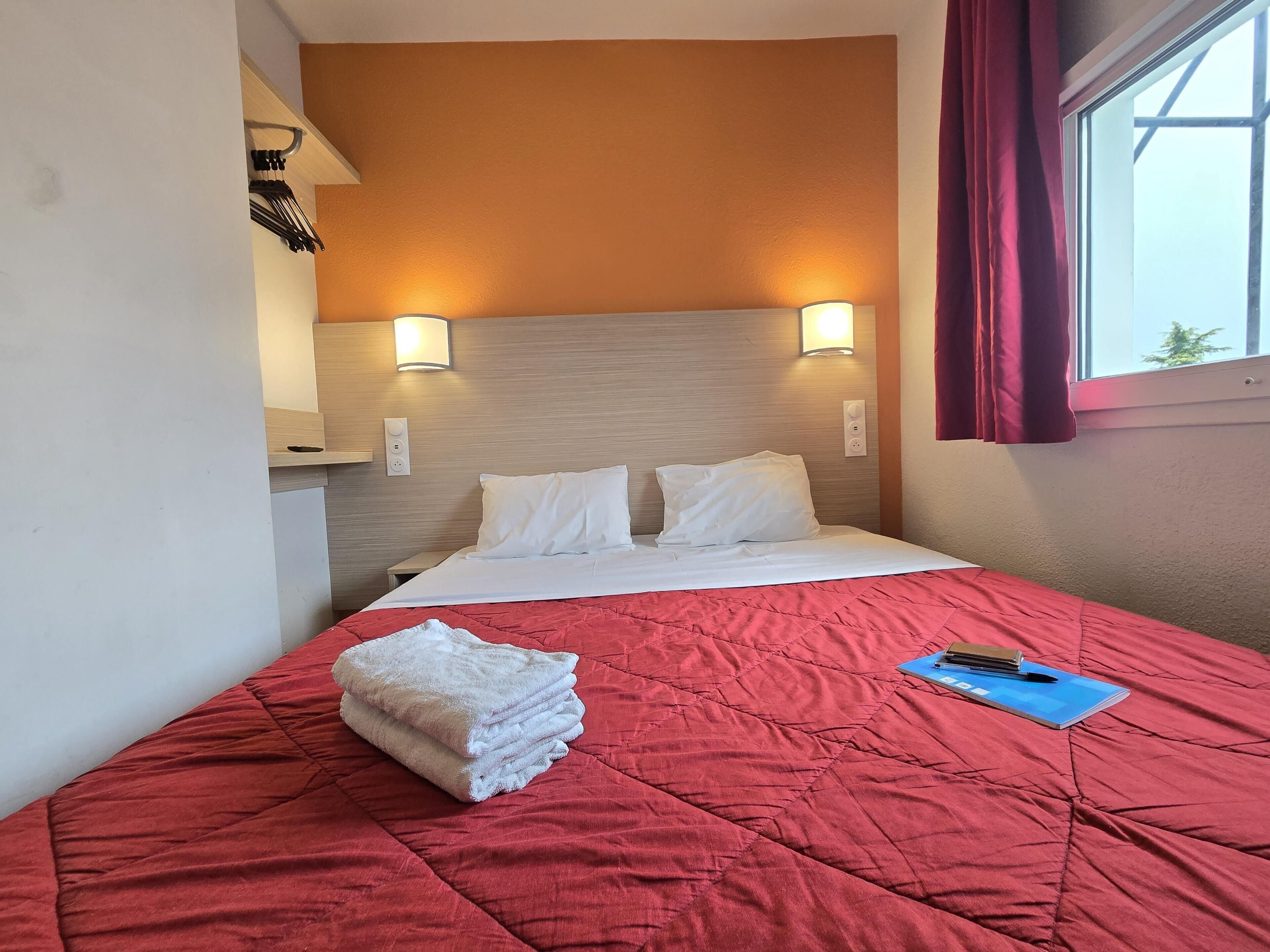 Habitación de hotel con cama, colcha acolchada roja, almohadas blancas, una libreta azul y una pila de toallas blancas.