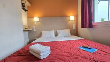Habitación de hotel con cama, colcha acolchada roja, almohadas blancas, una libreta azul y una pila de toallas blancas.