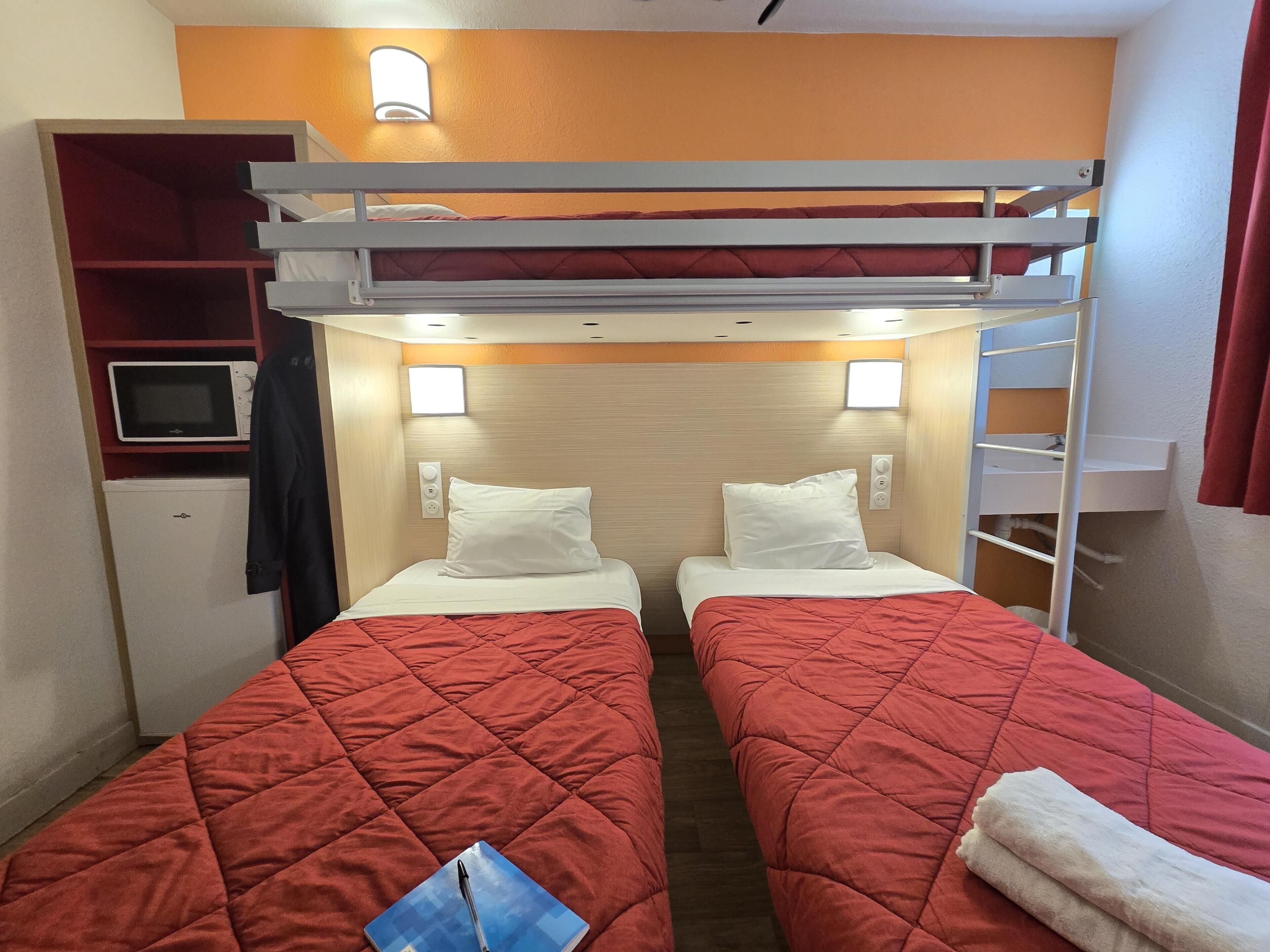 Une chambre avec deux lits, chacun recouvert d’une courtepointe rouge matelassée et agrémenté d’oreillers blancs. Une petite étagère, sur laquelle se trouvent une télévision et un four à micro-ondes, est placée à gauche.