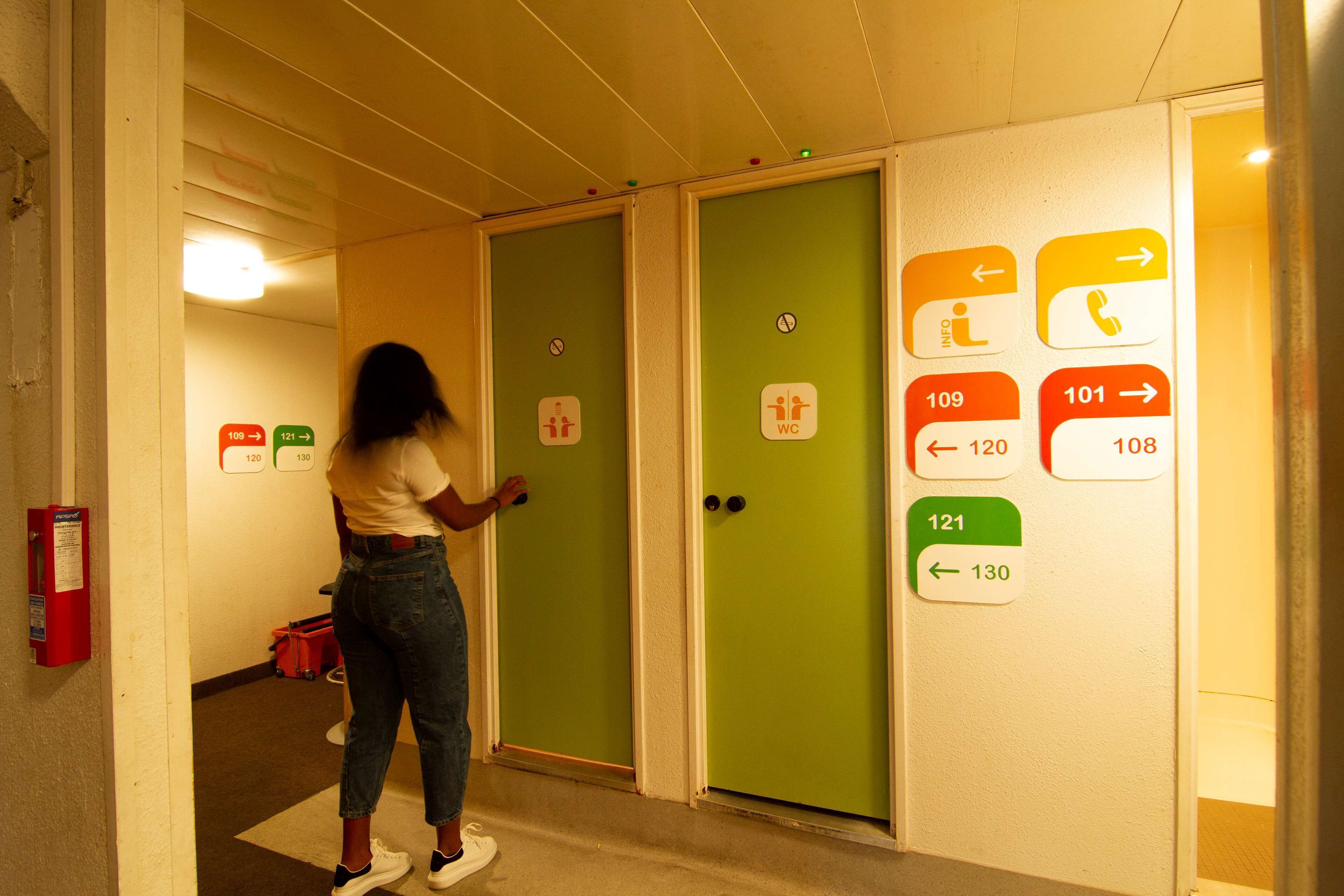 Un pasillo con puertas verdes, cada una con números y flechas indicando direcciones. Una persona está de pie cerca de las puertas, y un extintor es visible a la izquierda.