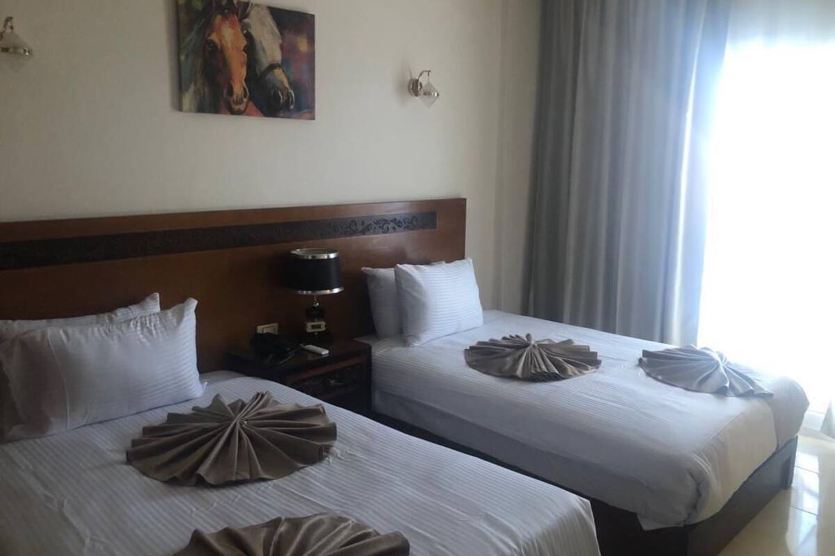 Une chambre d’hôtel avec deux lits, une tête de lit, un tableau, une table de chevet et une fenêtre avec des rideaux.