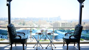 Un balcón con dos sillas tapizadas y una mesa de vidrio, con vistas a un paisaje urbano con edificios y cielo despejado.
