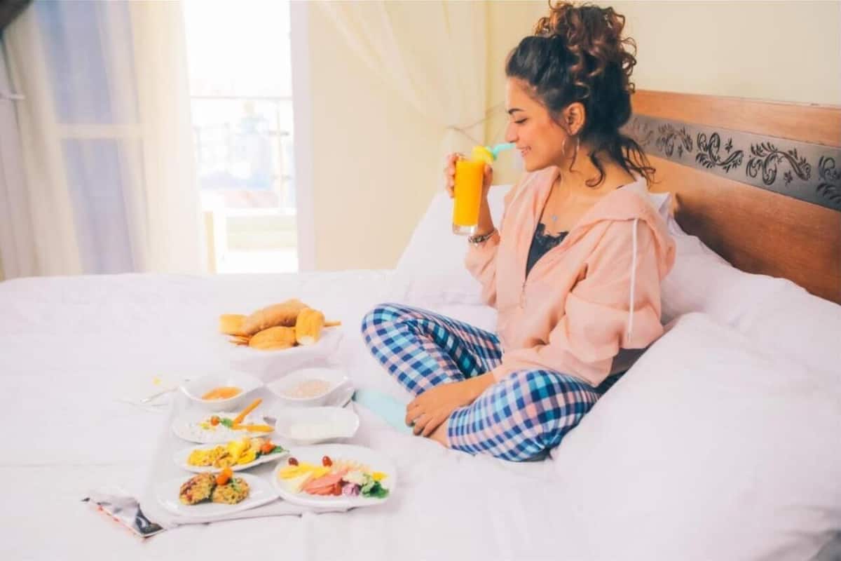 Une personne assise sur un lit, avec des aliments pour le petit-déjeuner et une boisson.