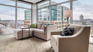 A modern living room with a sectional sofa, a small table, and a floor lamp. Large windows offer a view of a cityscape and a building with glass windows. - Cambria Hotel Louisville Downtown-Whiskey Row (Louisville)